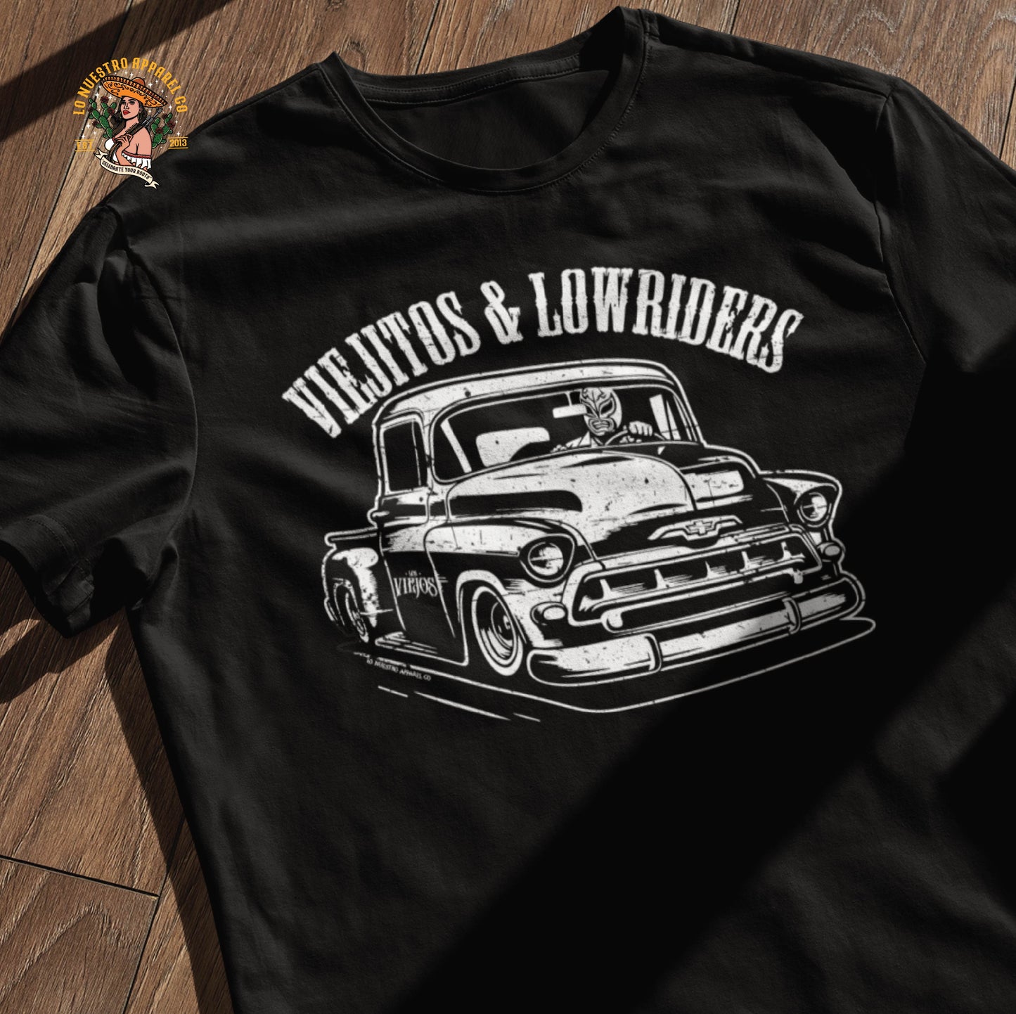 A flat lay of the Viejitos & Lowriders lowrider t-shirt on a wooden surface, showcasing the bold vintage design of a 1957 Chevy Stepside and luchador driver. A must-have in men’s lowrider shirts for those who rep Chicano clothing with style.