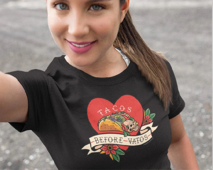 A black women's relaxed t-shirt with a colorful graphic of a taco and heart, with the text 'Tacos Before Vatos.