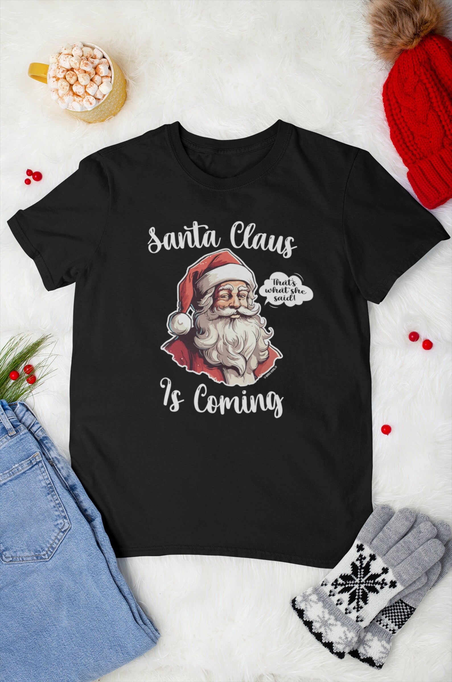 Flat lay of a "Santa Claus Is Coming" unisex shirt with festive winter accessories, including a red knit hat, cozy gloves, and a mug of hot chocolate, emphasizing the humorous holiday design.