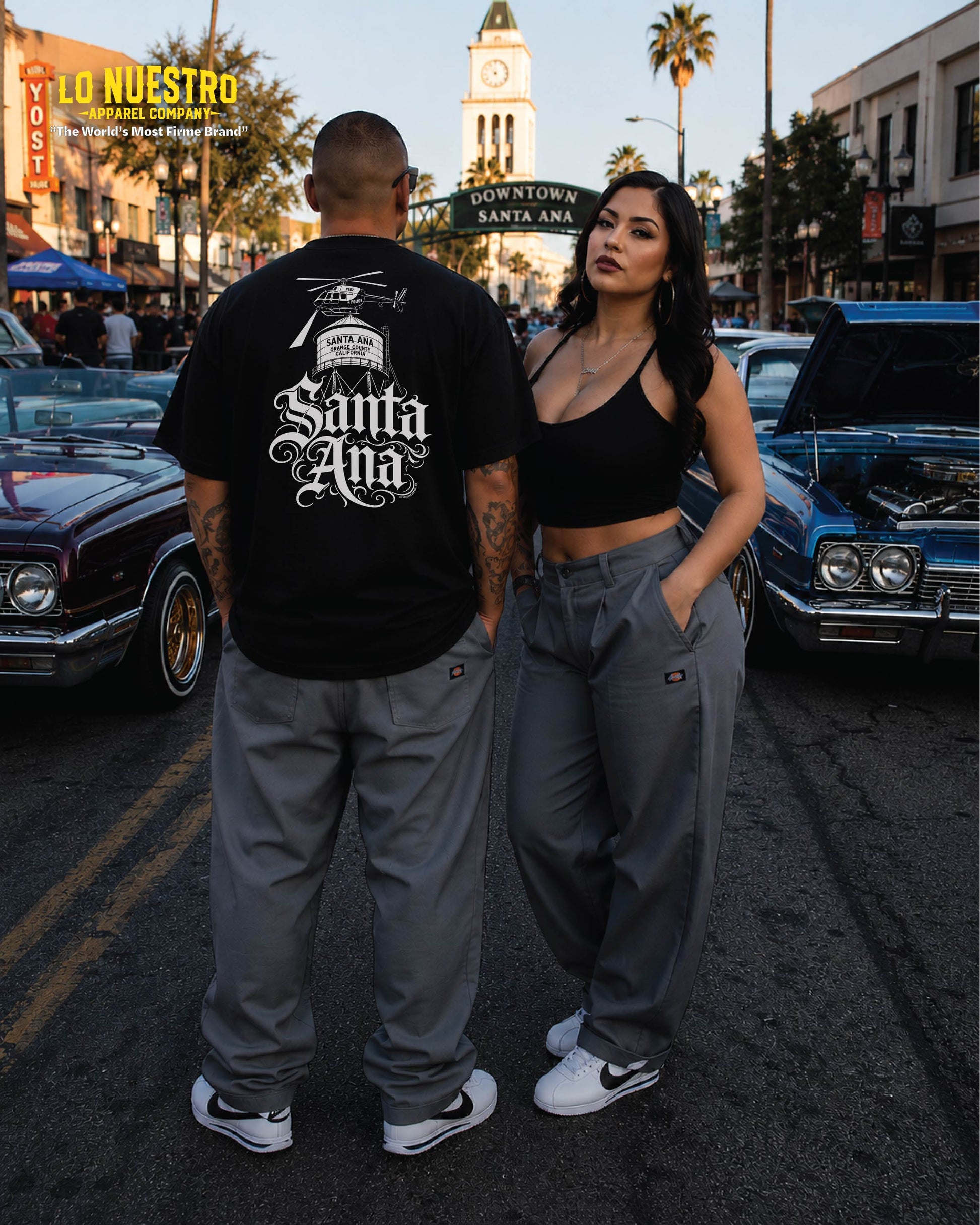 Man wearing the Santa Ana Old English T-shirt with back graphic featuring the Santa Ana water tower and helicopter standing with woman in downtown Santa Ana surrounded by lowriders.