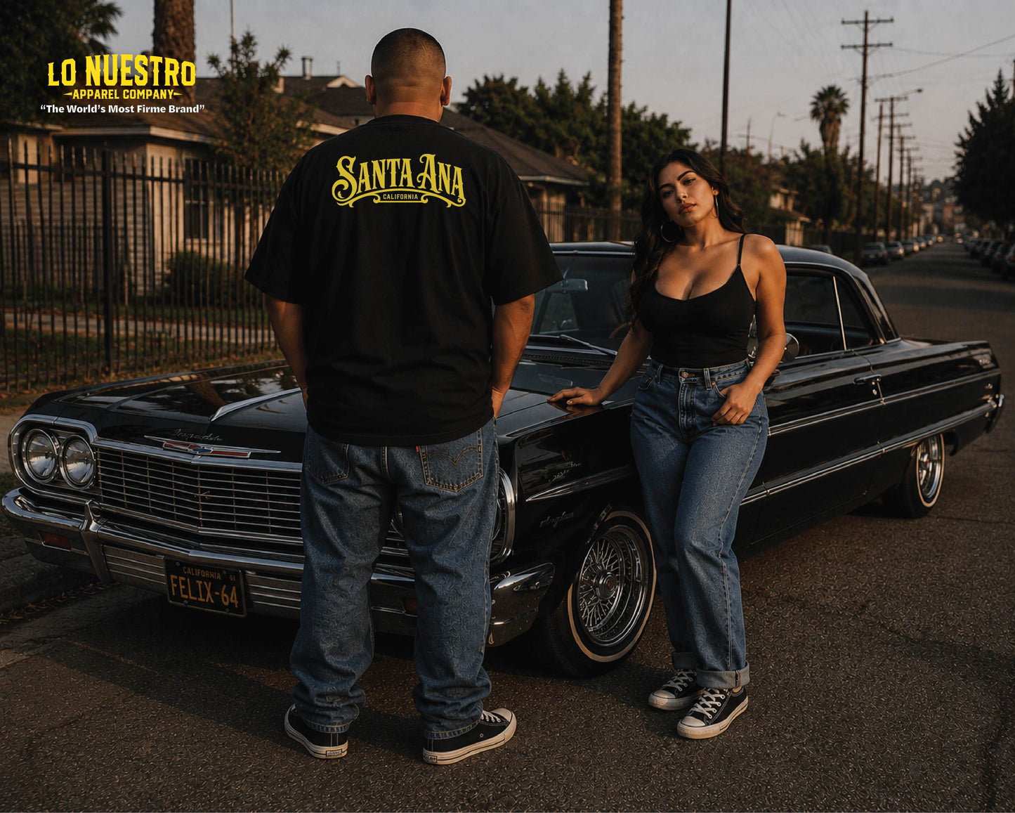 A lifestyle photo featuring a man and a woman standing next to a classic black car on a street during golden hour. The man is wearing a black t-shirt with 'Santa Ana' printed in yellow on the back, and the woman is wearing a black tank top and blue jeans.