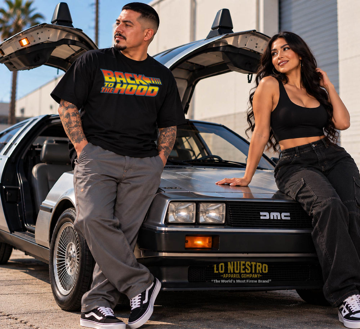 Chicano man wearing a black Back to the Hood T-shirt with a Latina woman posing beside a classic stainless steel sports car