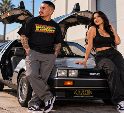 Chicano man wearing a black Back to the Hood T-shirt with a Latina woman posing beside a classic stainless steel sports car