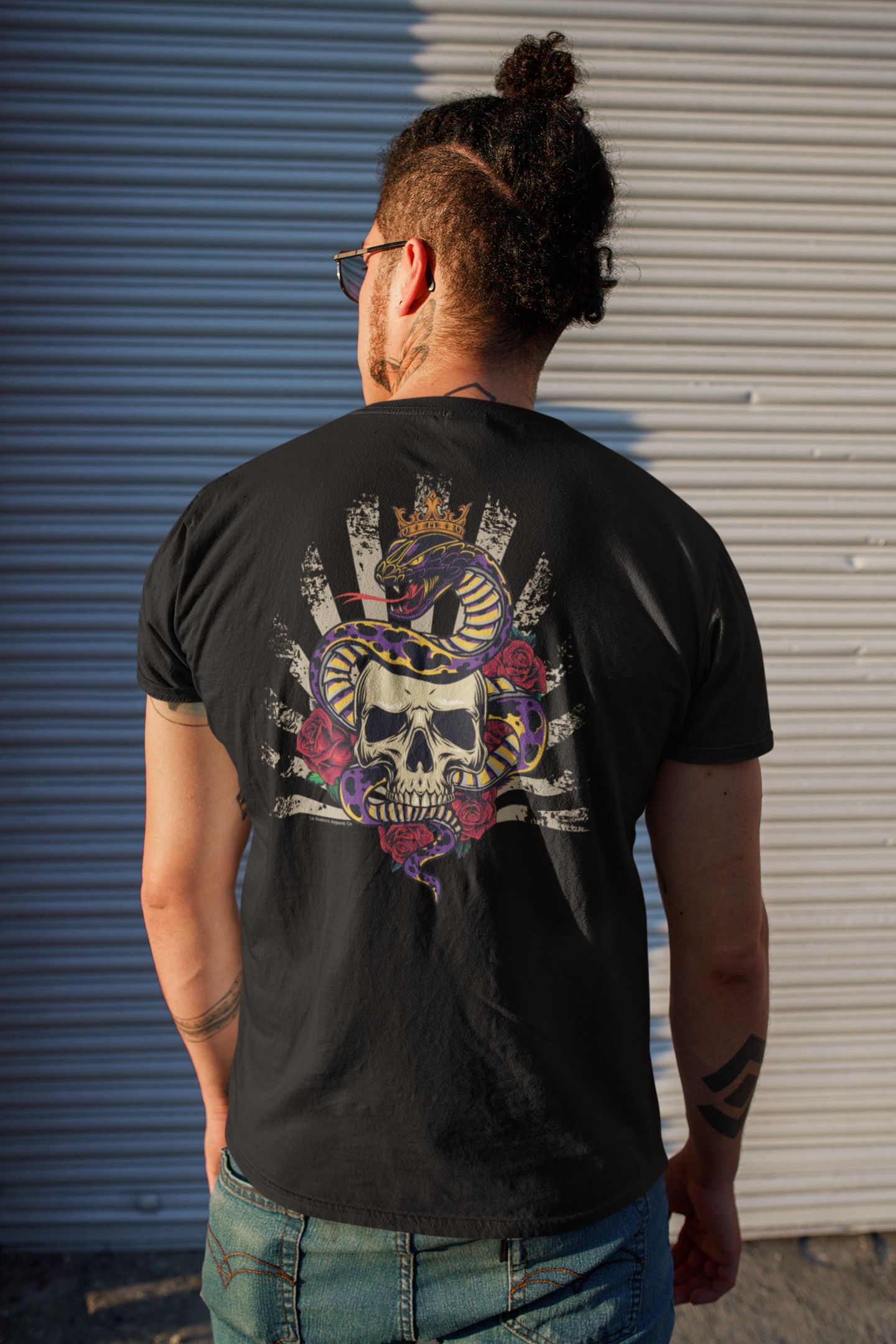 Man wearing the G.O.A.T. T-Shirt, showing the back graphic of a crowned snake wrapped around a skull, standing on the street with a bun hairstyle.