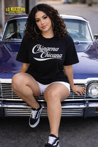 Latina woman sitting on a purple blue lowrider wearing a black Chingona Chicana t-shirt with script design, styled with hoop earrings and streetwear look