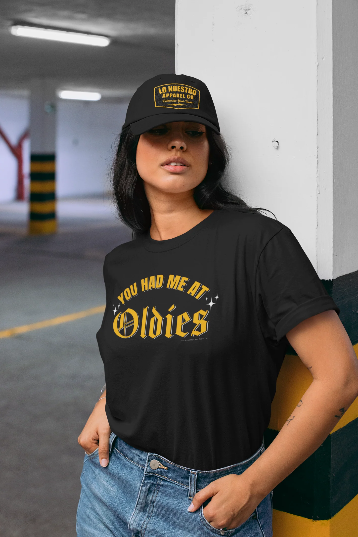 Woman wearing a black "You Had Me at Oldies" T-shirt with bold yellow old English lettering, standing in a parking lot and leaning against a wall, also wearing a black "Lo Nuestro Apparel Co." hat.