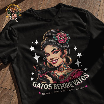 Black t-shirt laid flat on a wooden surface featuring the Gatos Before Vatos Tattoo T-Shirt design with a playful cat illustration. Mexican  clothing.