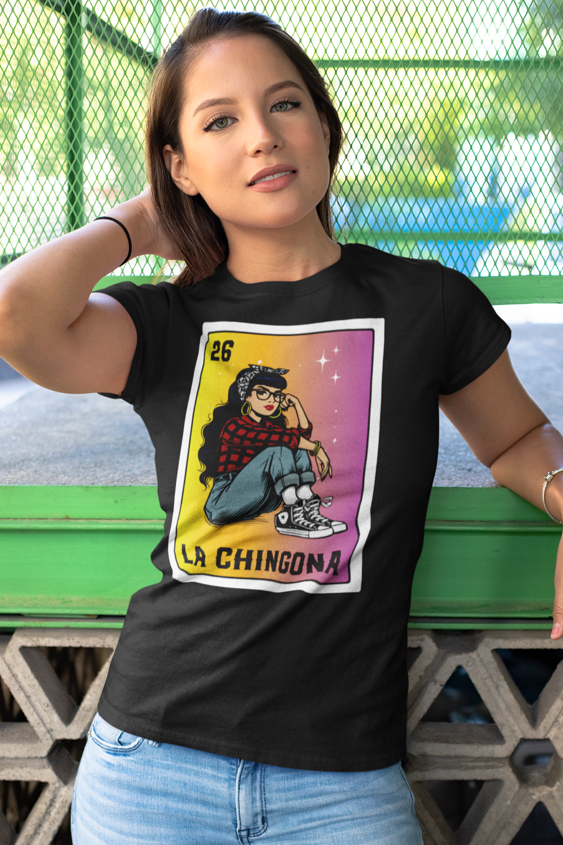 A young woman leaning against a green bench, wearing a black "La Chingona" Lotería-style t-shirt, featuring a Chicana in a red plaid shirt, jeans, and Converse sneakers, set against a bold yellow-to-pink gradient.