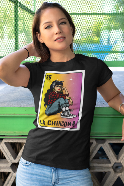 A young woman leaning against a green bench, wearing a black "La Chingona" Lotería-style t-shirt, featuring a Chicana in a red plaid shirt, jeans, and Converse sneakers, set against a bold yellow-to-pink gradient.