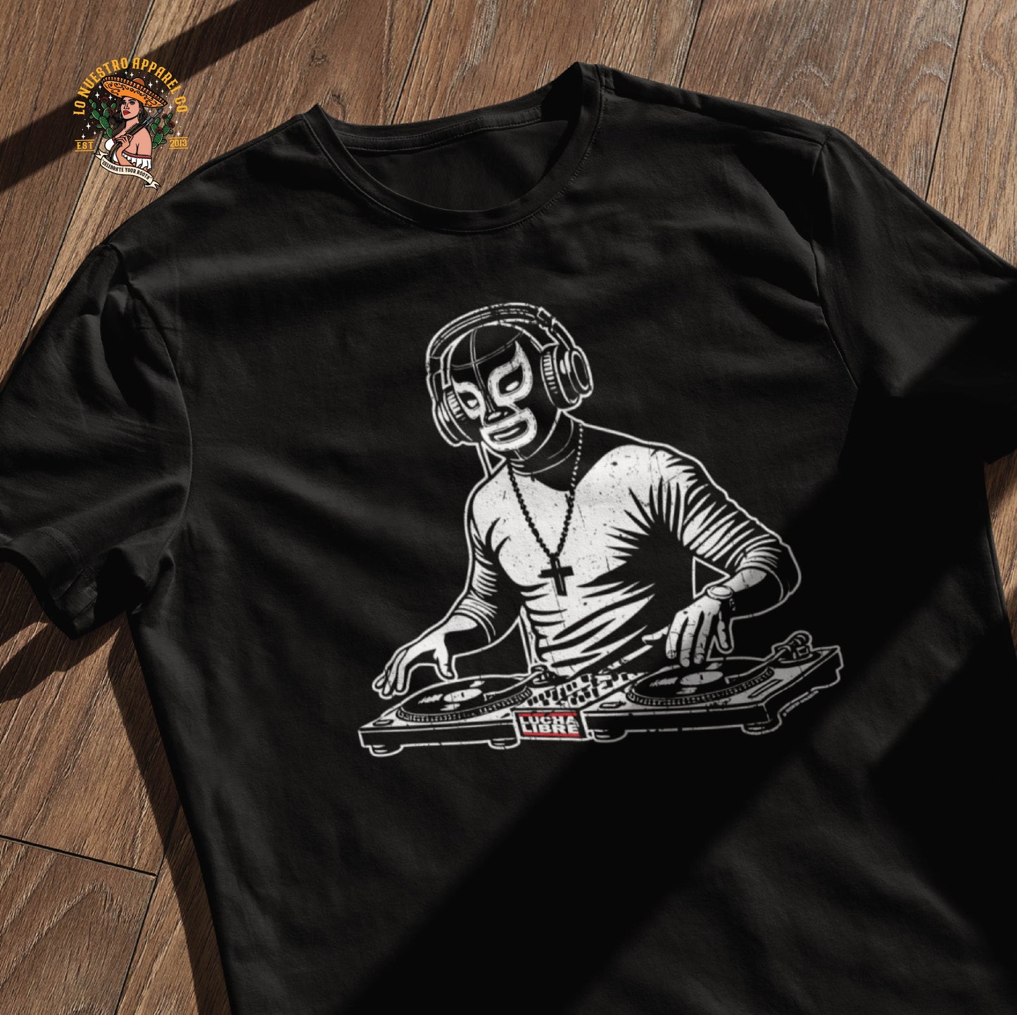 A black Luchador Shirt laid on a wooden surface, featuring a masked DJ spinning records on turntables. This Funny Mexican Shirt blends Chicano clothing with music culture, making it a must-have for those who rep both beats and lucha style.