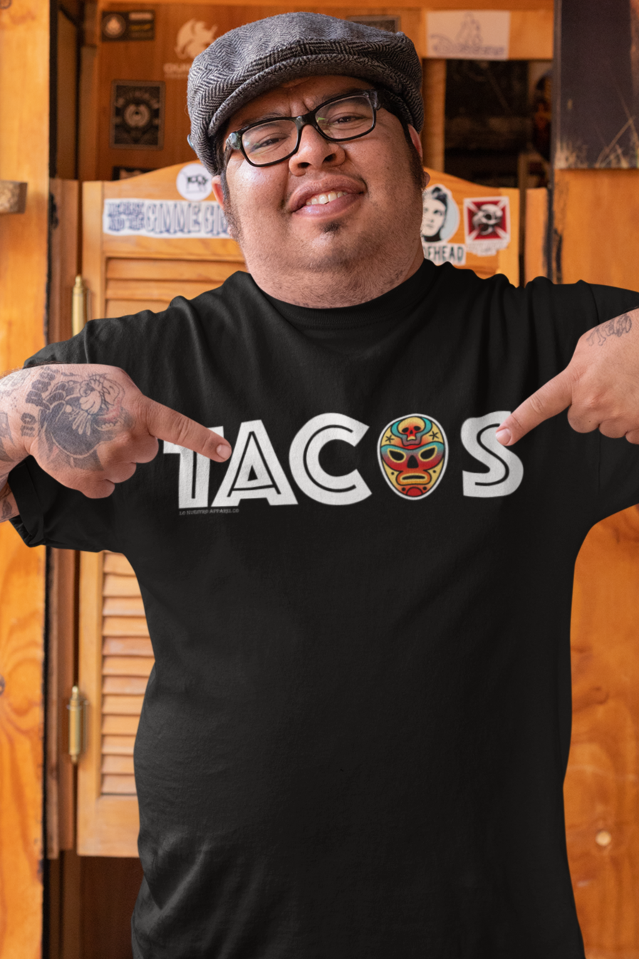 Man wearing a black Luchador Tacos T-shirt, pointing at the bold "TACOS" text with a colorful luchador mask design. Mexican food and wrestling-inspired streetwear.