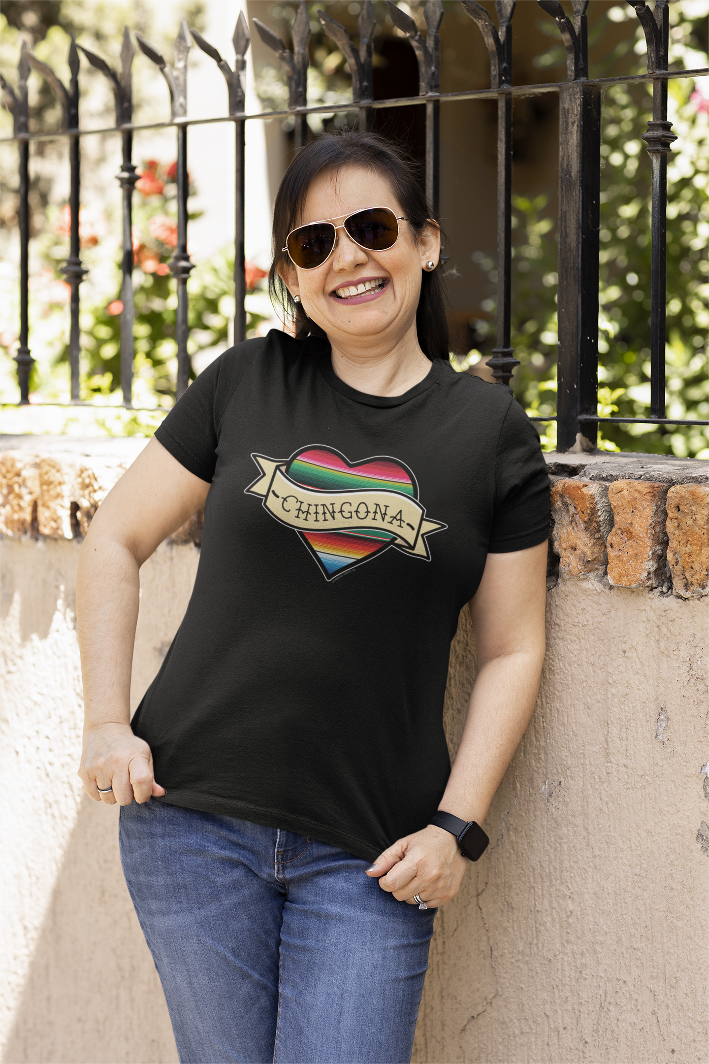 Smiling woman wearing a black T-shirt with a serape heart and 'Chingona' text, representing Chicana pride.