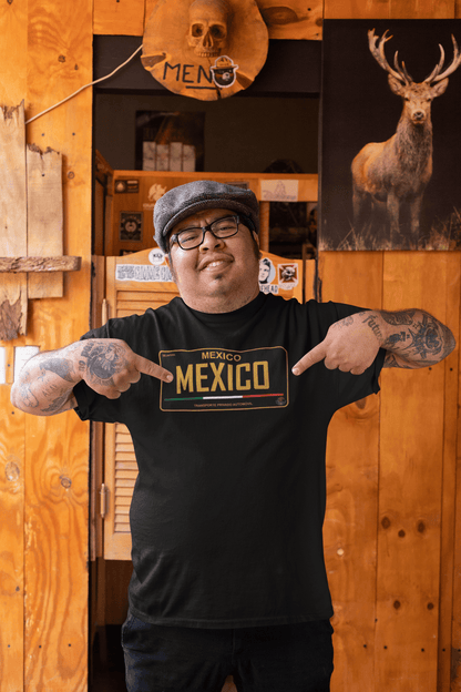 A man in a rustic bar wearing a black t-shirt featuring a vintage-style Mexico license plate graphic in bold gold and tricolor accents.