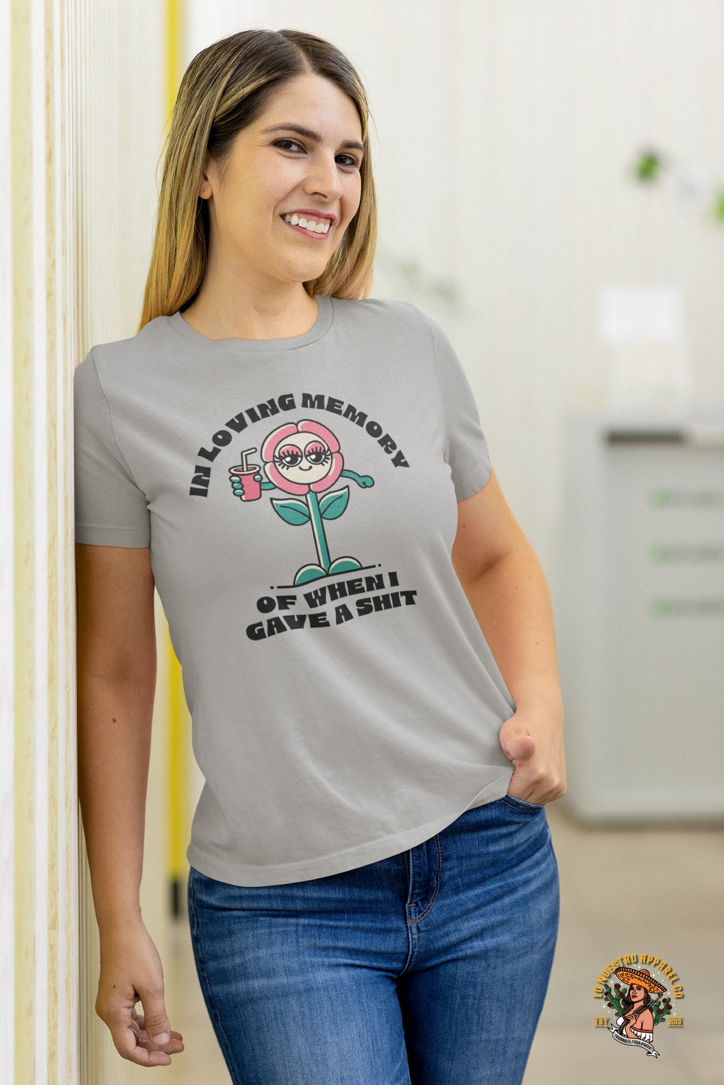 Smiling woman wearing the 'In Loving Memory of When I Gave a Sh*t' T-Shirt in Athletic Heather, featuring a quirky cartoon flower graphic.