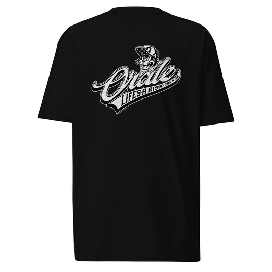 Flat-lay of a black 'Órale Life’s a Risk Carnal' T-shirt featuring a bold baseball-style script with an angry cat graphic in a bandana. Chicano streetwear with a lowrider and barrio-inspired design.