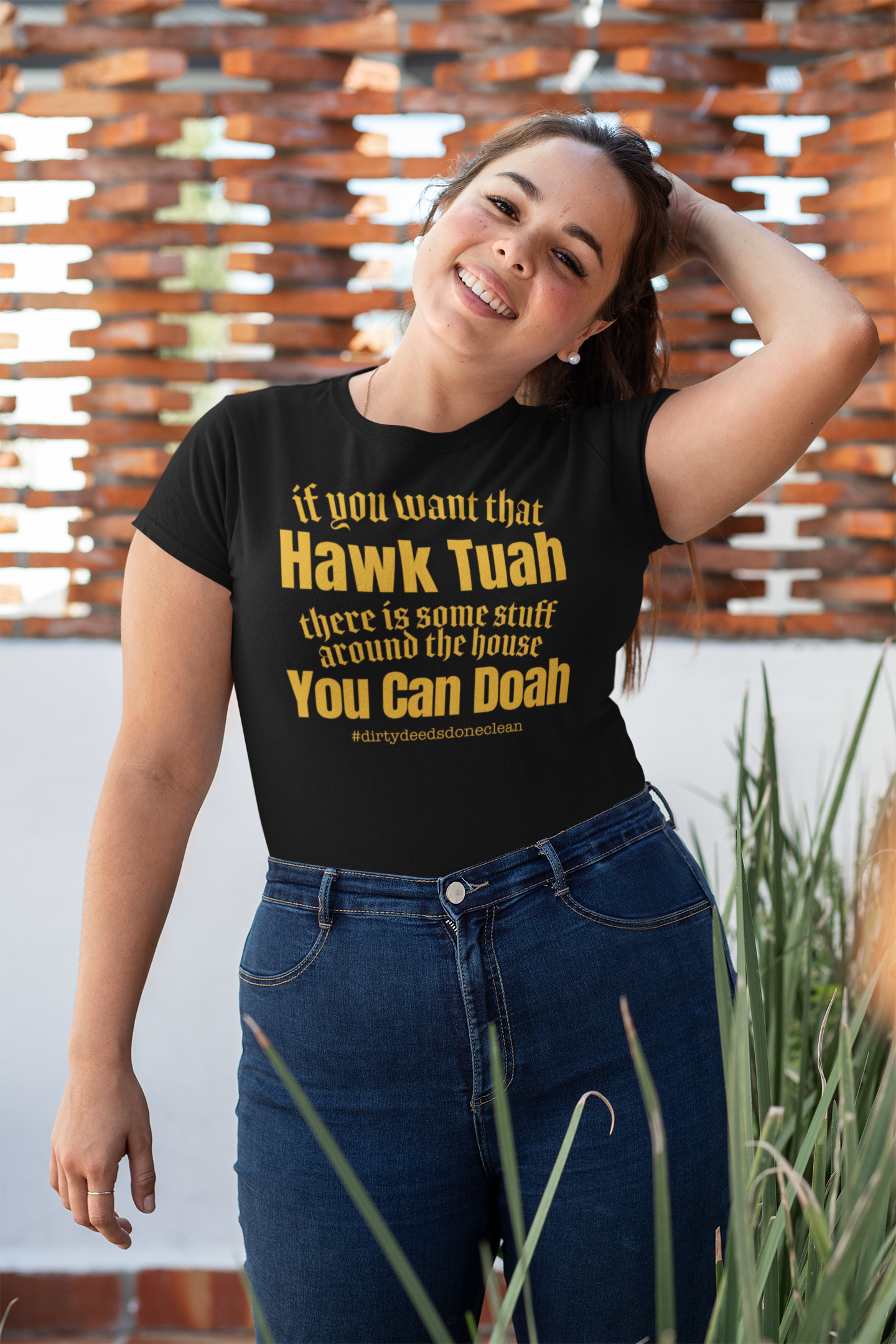 A smiling woman wearing a black relaxed t-shirt with the text: 'If you want that Hawk Tuah, there is some stuff around the house you can doah' in bold yellow, standing in a cozy living room.