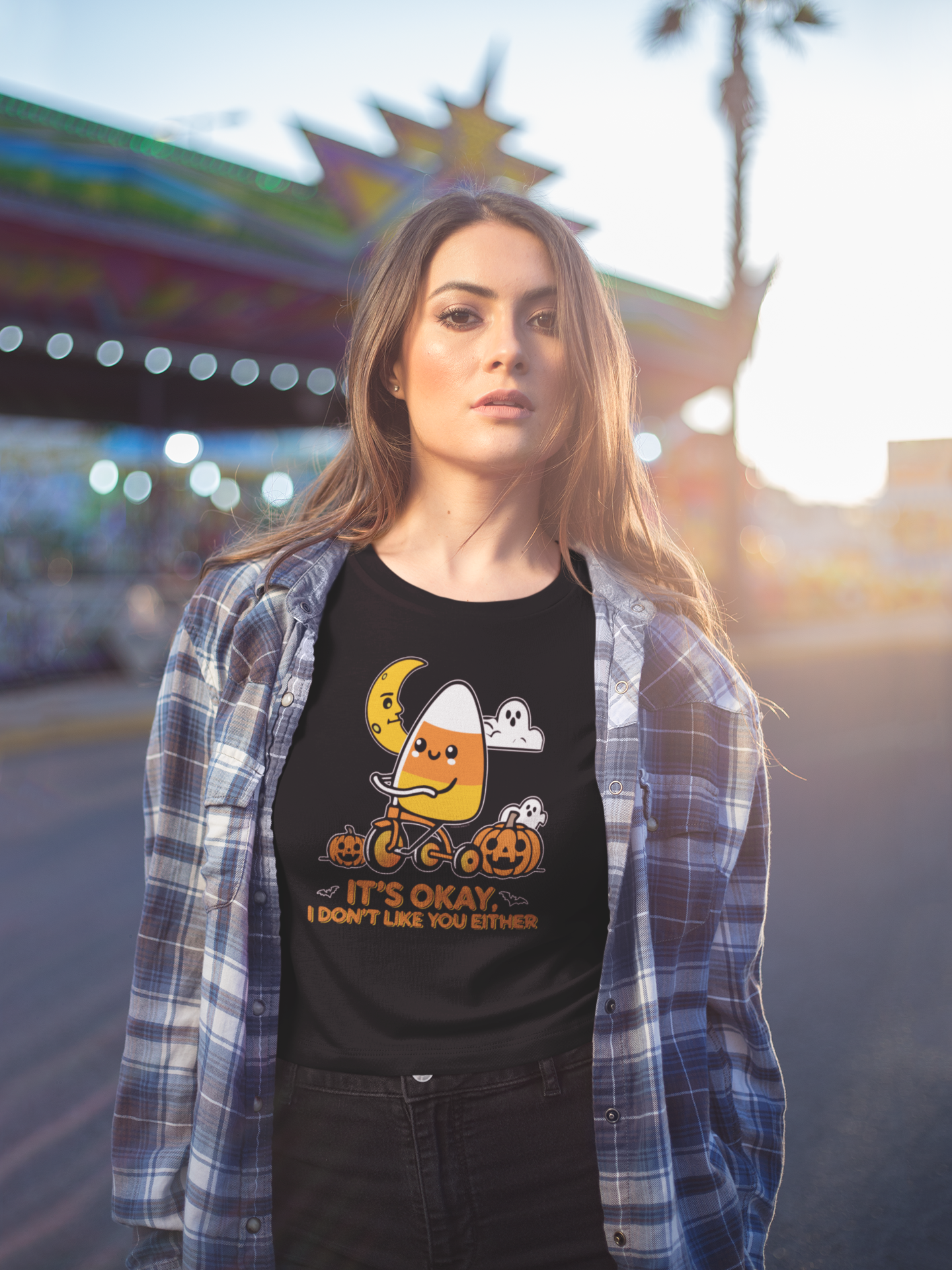 A woman wearing a black t-shirt featuring a cute candy corn character riding a tricycle, accompanied by pumpkins and ghosts under a crescent moon. The t-shirt reads 'It’s okay, I don’t like you either,' combining playful humor with a Halloween theme. She stands outdoors in a casual, laid-back look, with a plaid shirt layered over the tee, against the backdrop of a carnival.