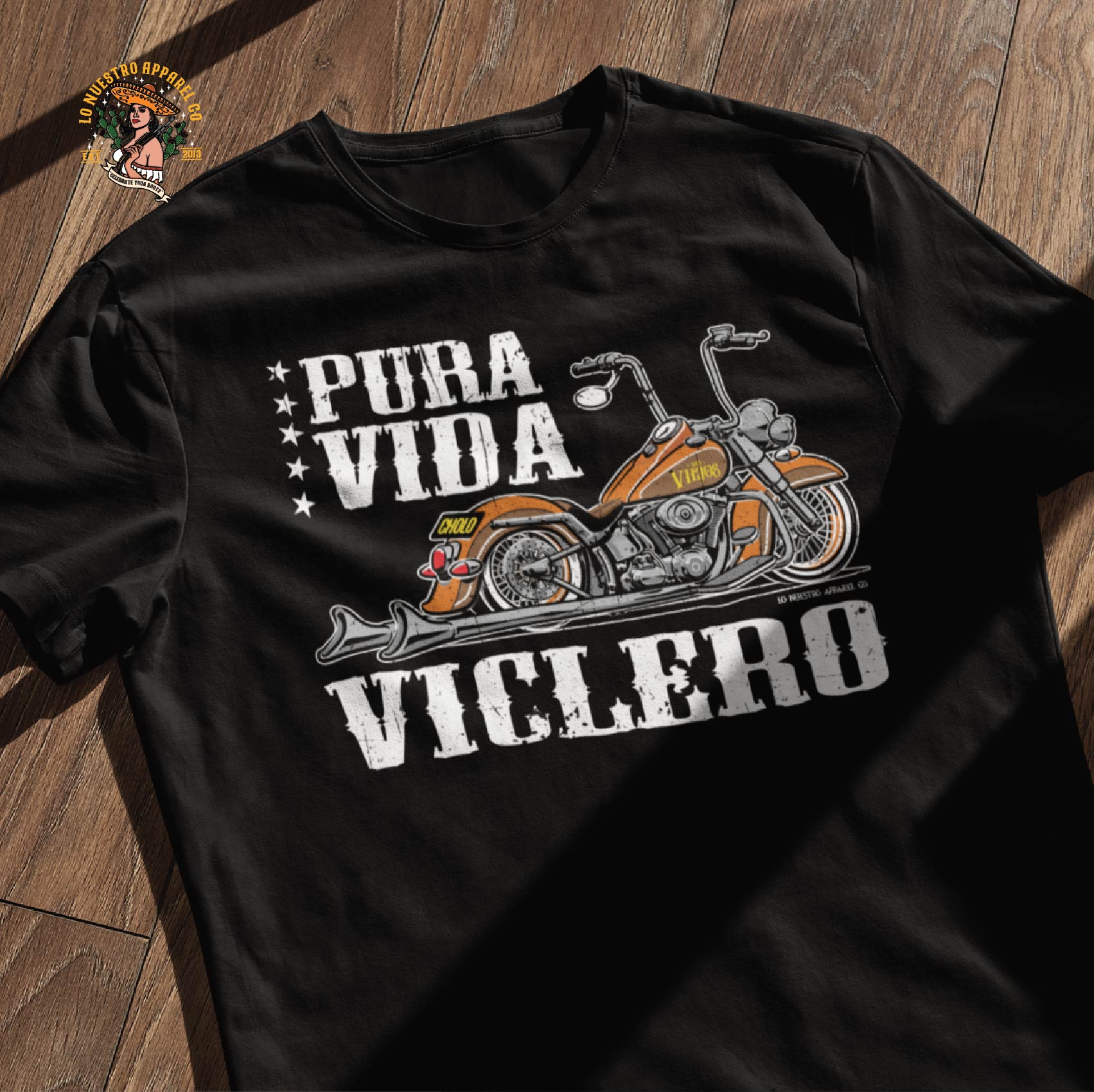 The Pura Vida Viclero T-Shirt displayed on a wooden surface, showcasing its detailed motorcycle design. This biker t-shirt blends Chicano clothing with street culture, making it a must-have for riders and those who live by the road. A perfect addition to any collection of funny Mexican shirts with a tough, vintage-inspired look.