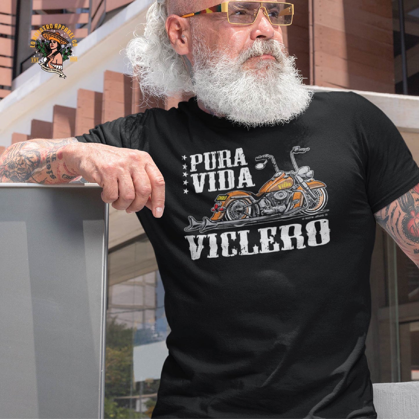 A stylish older man with a white beard wearing the Pura Vida Viclero T-Shirt, a Chicano clothing piece designed for bikers. This biker t-shirt features a motorcycle graphic and represents the Chicano lifestyle with firme style. Perfect for fans of funny Mexican shirts and streetwear with attitude.