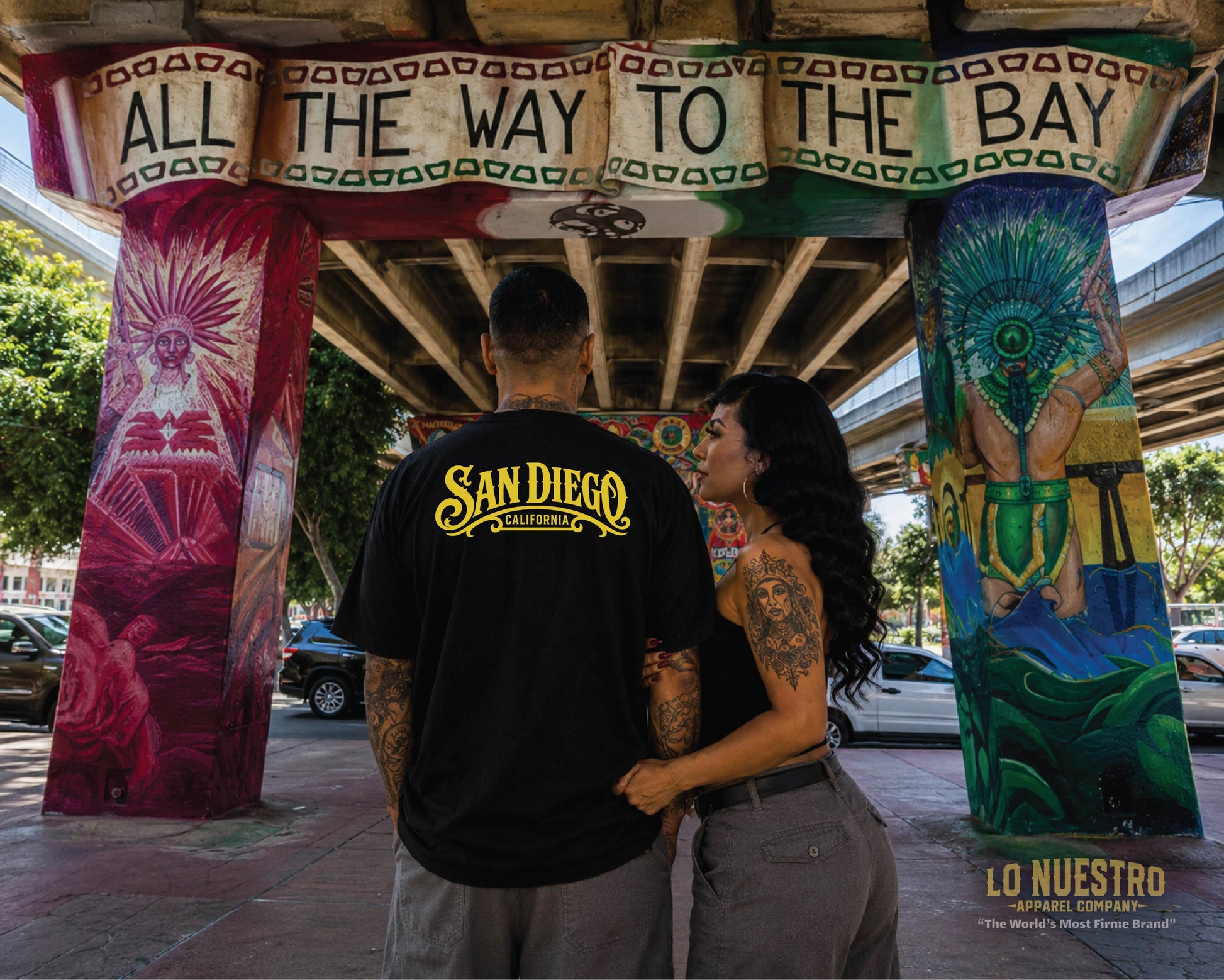 Back view of the San Diego Lowrider Plaque T-shirt worn beneath the iconic "All the Way to the Bay" mural in Chicano Park, San Diego.