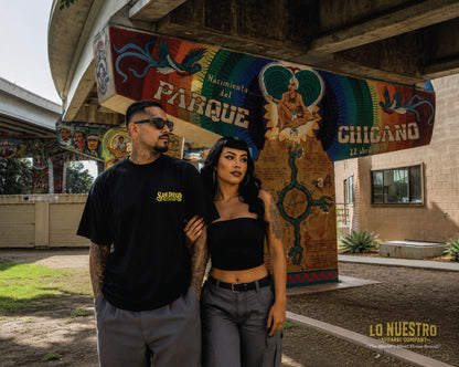 Man and woman wearing the San Diego Lowrider Plaque T-shirt in Chicano Park with the iconic murals beneath the Coronado Bridge in San Diego.