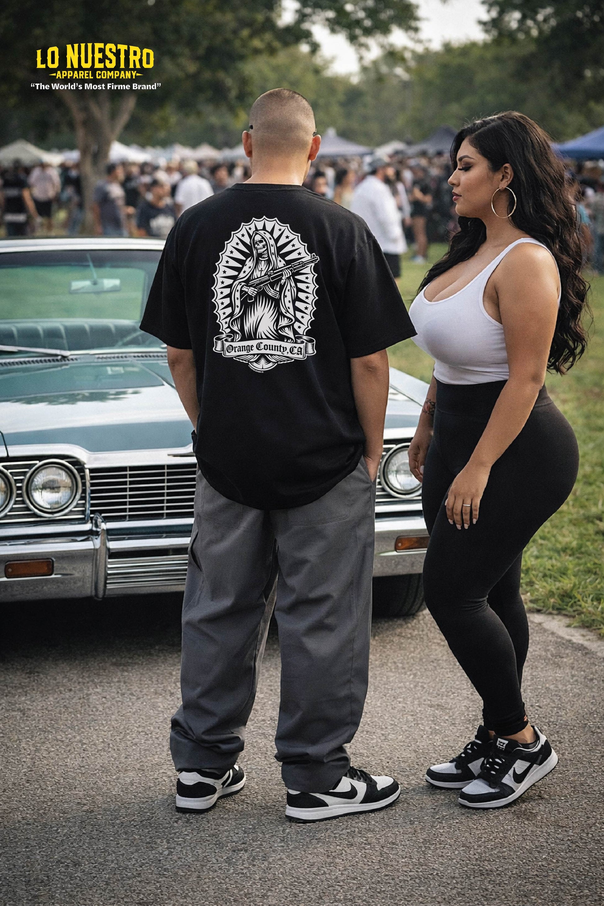 man wearing black Santa Muerte skull t-shirt standing in front of lowrider with woman at car show in park