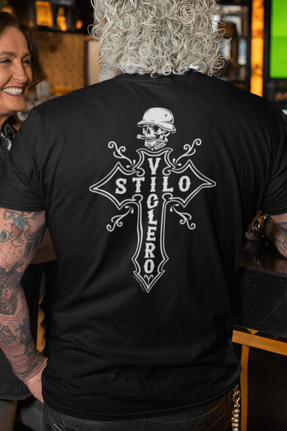 Back view of a black Stilo Viclero T-Shirt featuring a Chicano biker design with a cross, skull, and bold lettering, worn by a tattooed older man in a bar setting.