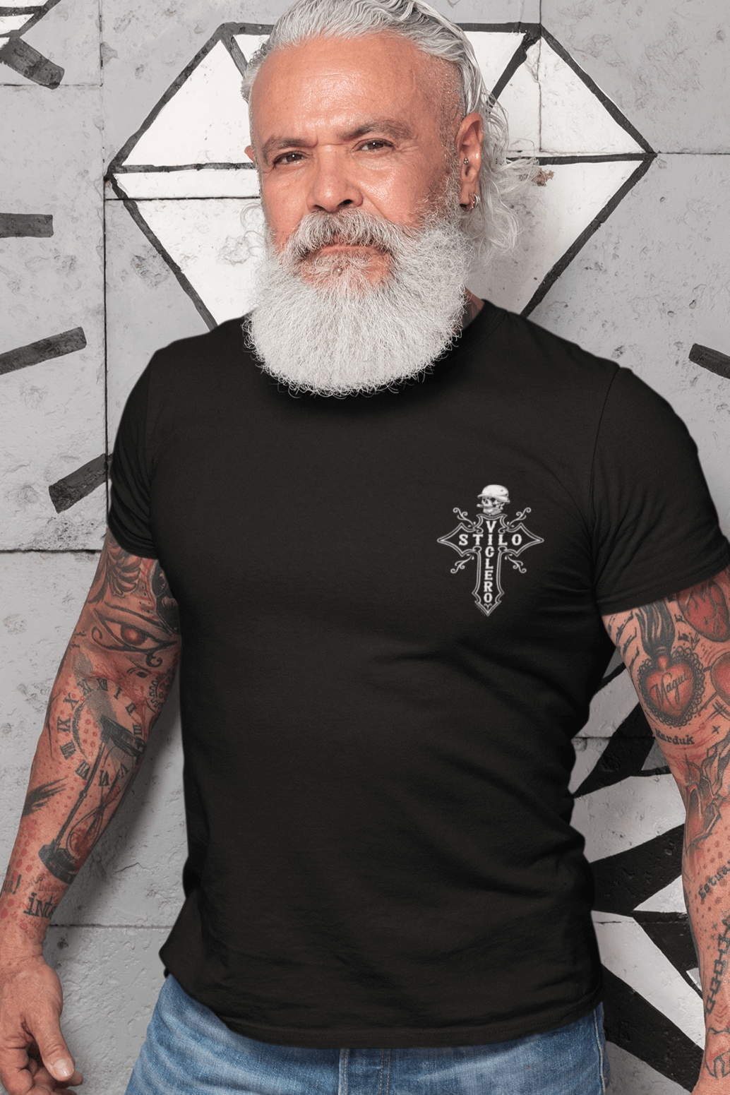 Tattooed older man with a white beard wearing a black Stilo Viclero T-Shirt, featuring a Chicano biker-inspired design with a skull and cross graphic on the left chest. Styled with blue jeans, standing against a graffiti-style background.