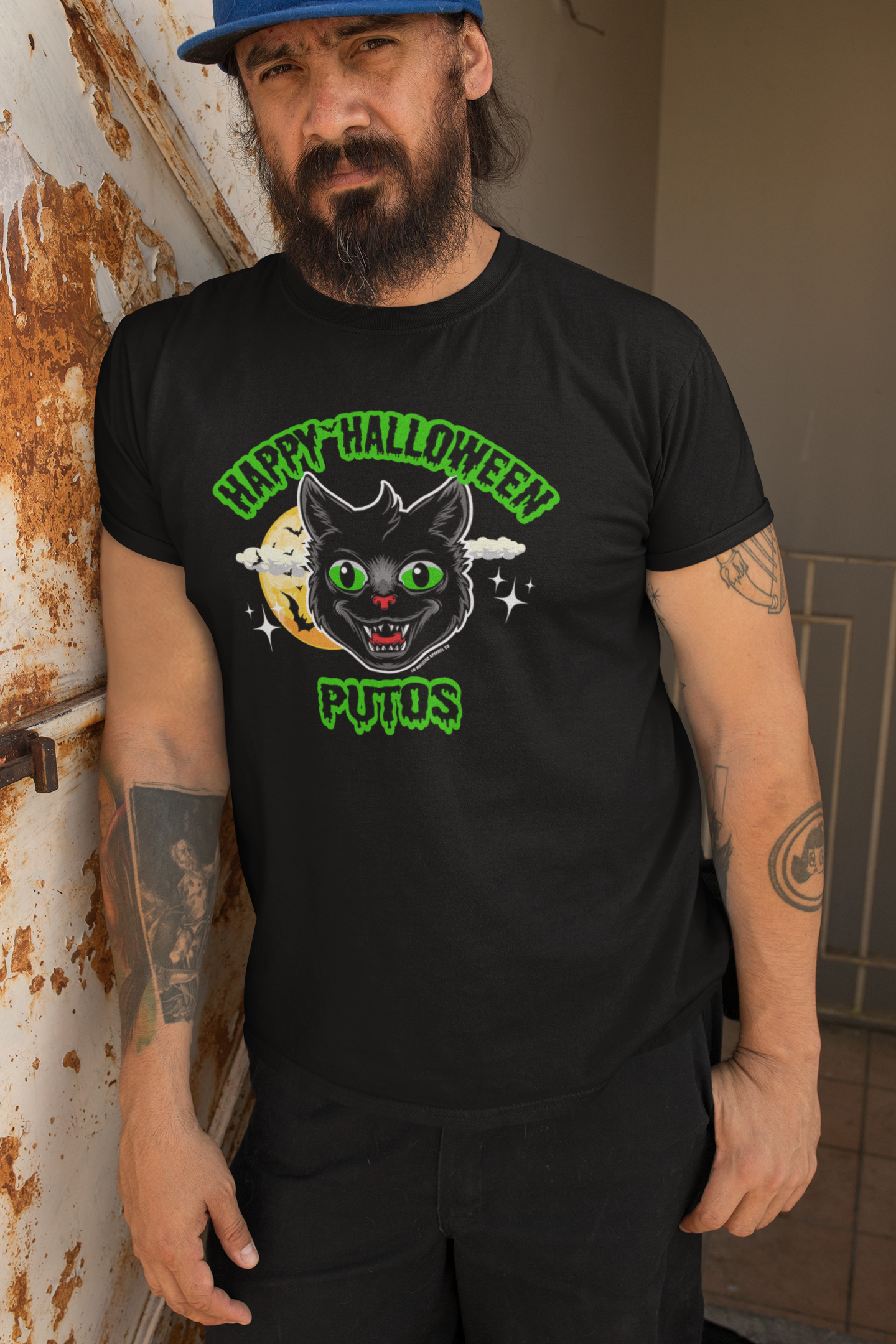 Man wearing Happy Halloween Putos Men's T-Shirt with black cat graphic leaning against a rusty wall – Funny Halloween T-Shirt, Halloween Graphic Tee, Stylish Halloween Wear.