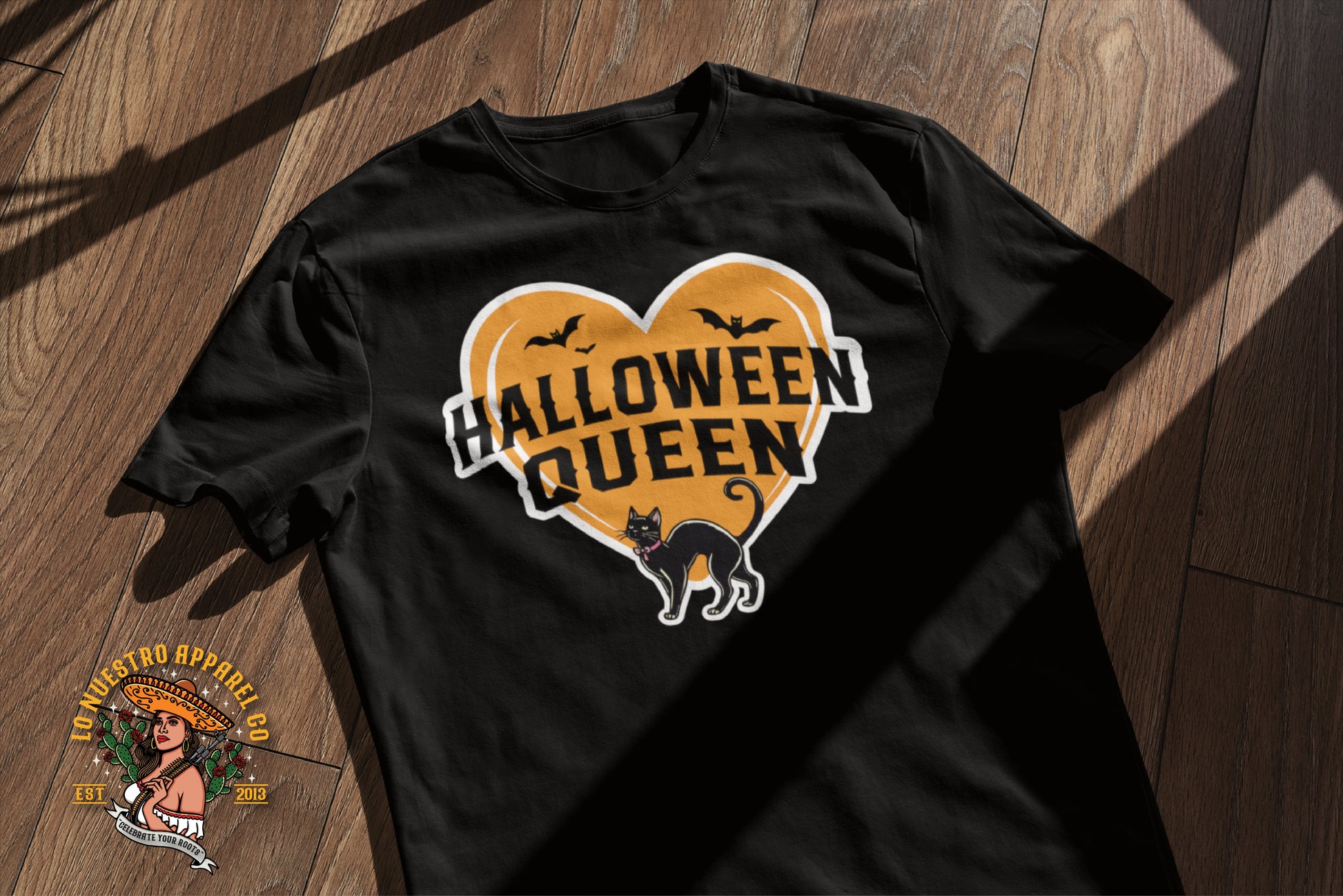 Halloween Queen' Women’s T-Shirt in Black, laid flat on a wooden surface, showcasing its vibrant orange heart graphic with a black cat and flying bats design.