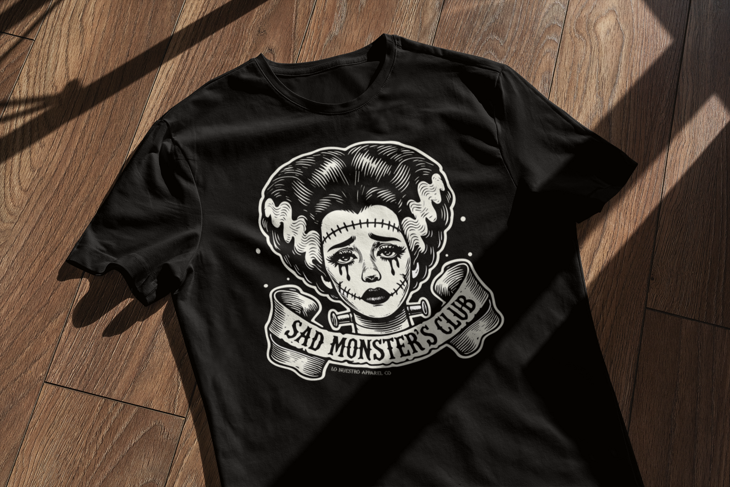 Black t-shirt with a retro tattoo-style 'Sad Monster's Club' Bride of Frankenstein design, laid flat on a wooden surface with window shadows adding a dramatic effect.