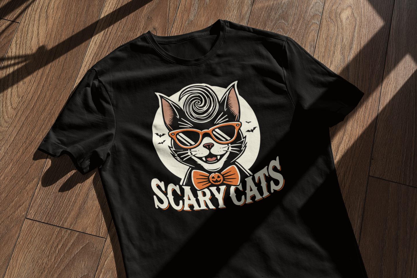 Folded black t-shirt with a retro 'Scary Cats' design featuring a cool cat in a greaser style and jack-o-lantern bow tie, placed on a wooden surface.