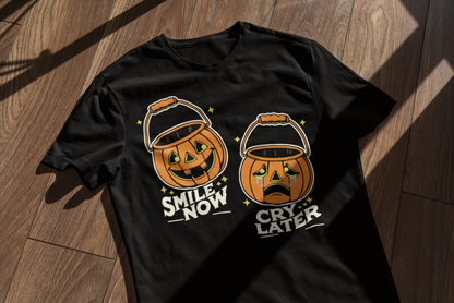 A black t-shirt with the 'Smile Now, Cry Later' jack-o'-lantern design, laid flat on a wooden surface with window shadows cast across it. The retro Halloween design is clearly displayed.