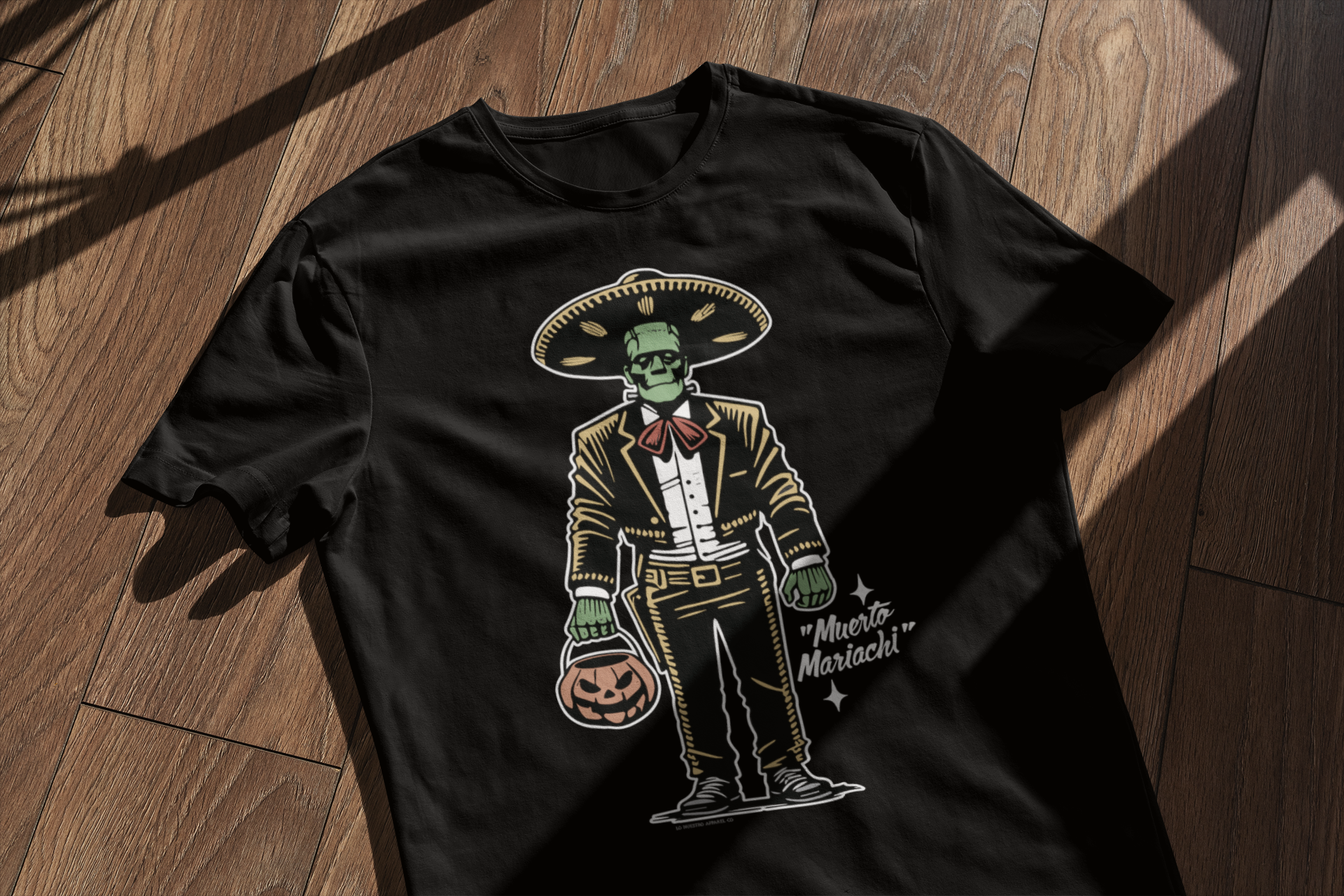 Black t-shirt laid on a wooden surface, featuring the 'Muerto Mariachi' design with a green, zombie-themed mariachi character holding a pumpkin bucket, in retro Halloween style.