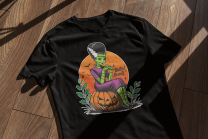 Black t-shirt featuring a retro Halloween design of a green-skinned bride sitting on a jack-o'-lantern with the phrase 'Bolted Beauty' in a spooky font. The shirt is laid flat on a wooden surface with light shadows.