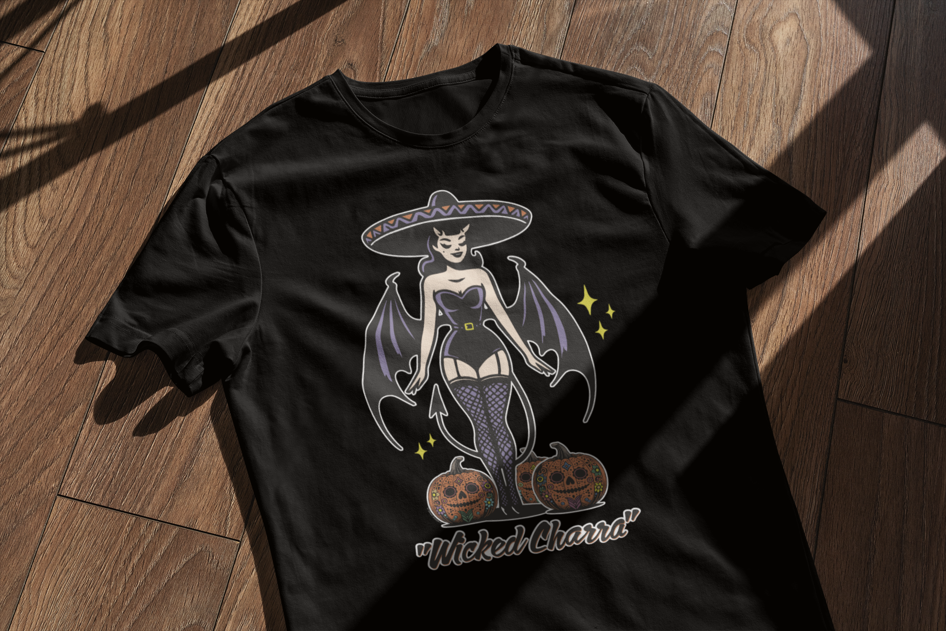 Black t-shirt laid flat on a wooden surface, featuring a 'Wicked Charra' design with a retro charra, devil wings, sombrero, and jack-o-lanterns, merging Halloween and Día de los Muertos elements.