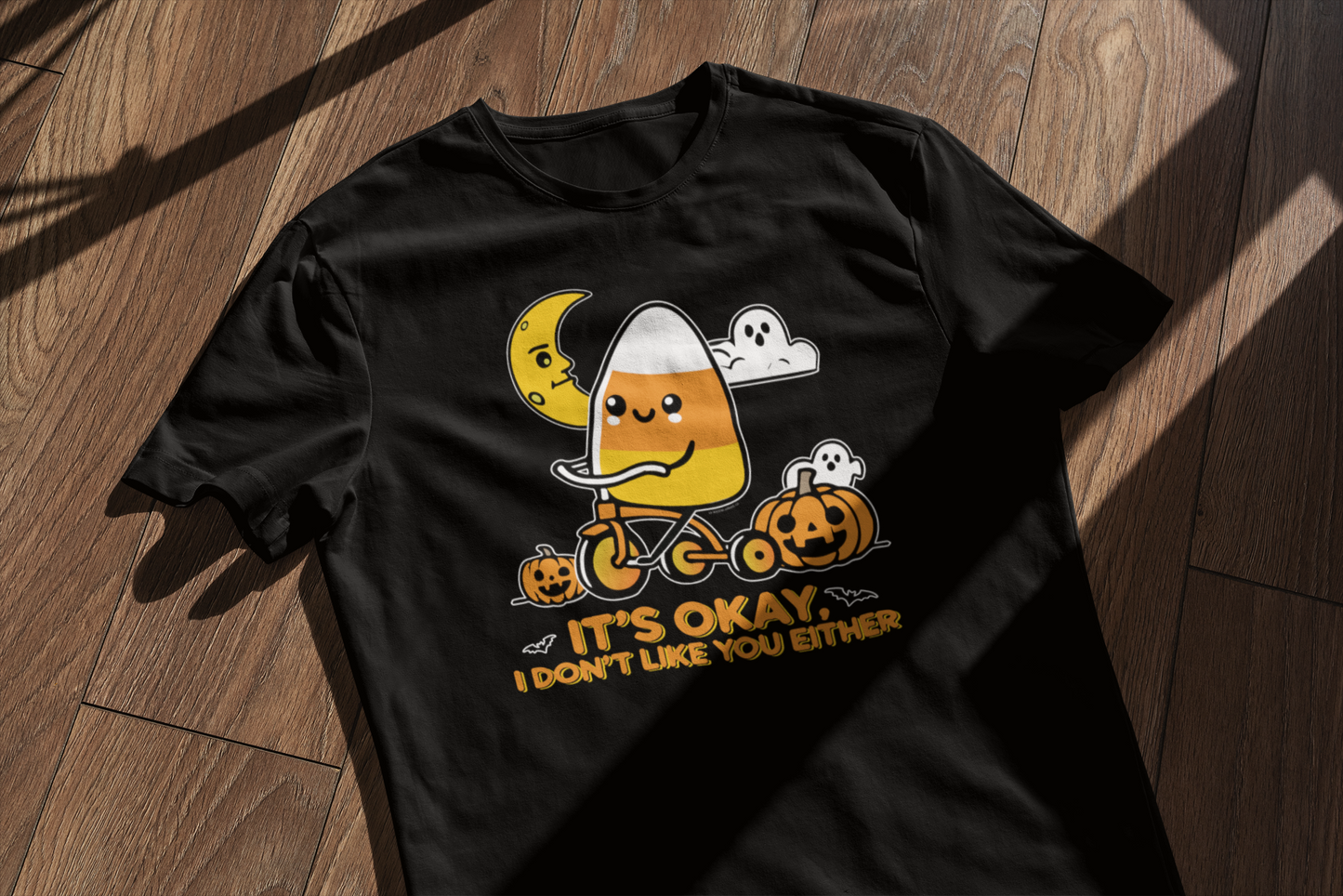 A black t-shirt laid on a wooden floor with a cute Halloween-themed design featuring a candy corn character riding a tricycle, surrounded by pumpkins, a crescent moon, ghosts, and the text "It's okay, I don't like you either" at the bottom.