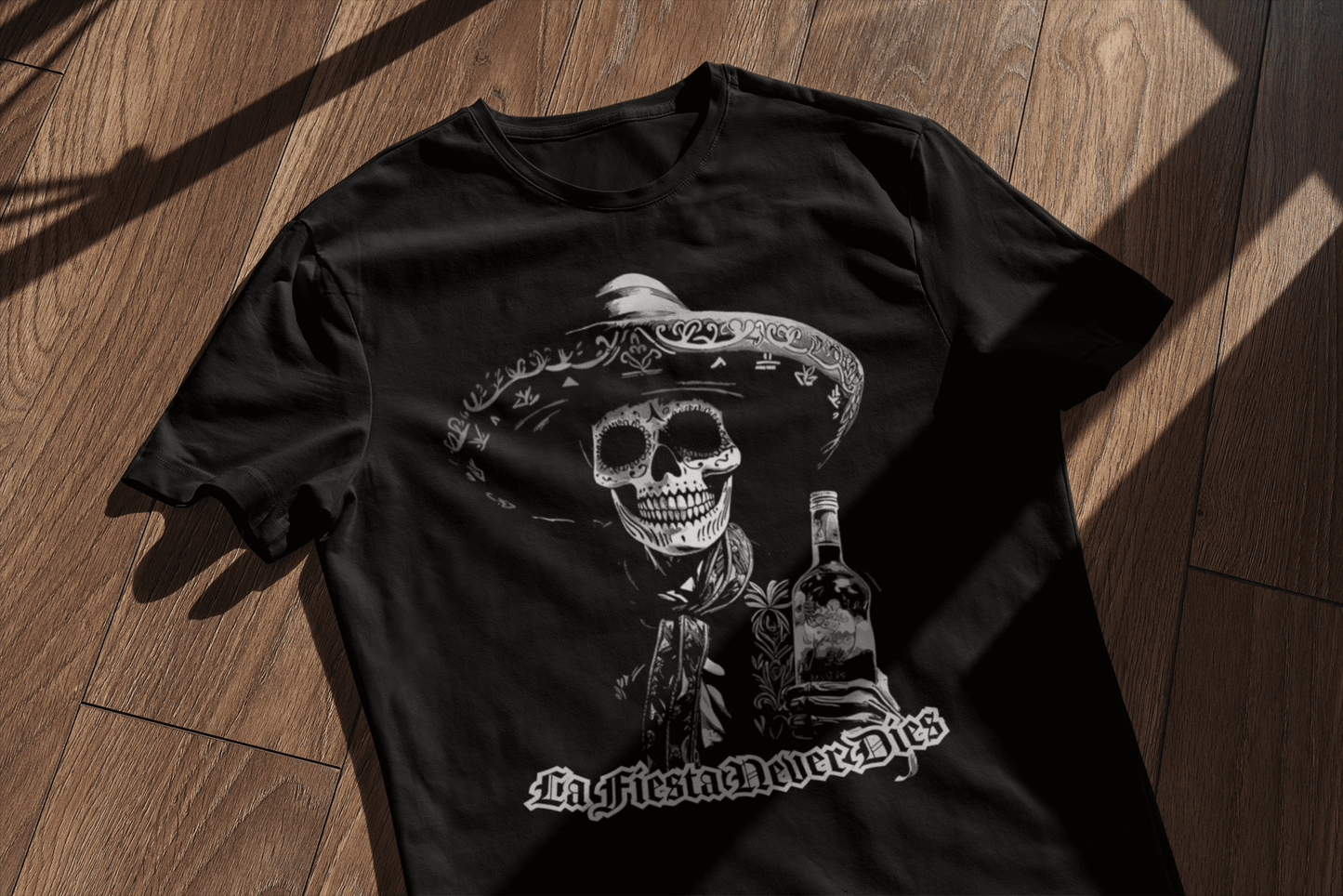 Black t-shirt laid on a wooden surface, featuring a bold 'La Fiesta Never Dies' Dia de los Muertos design with a charro skeleton holding a bottle.