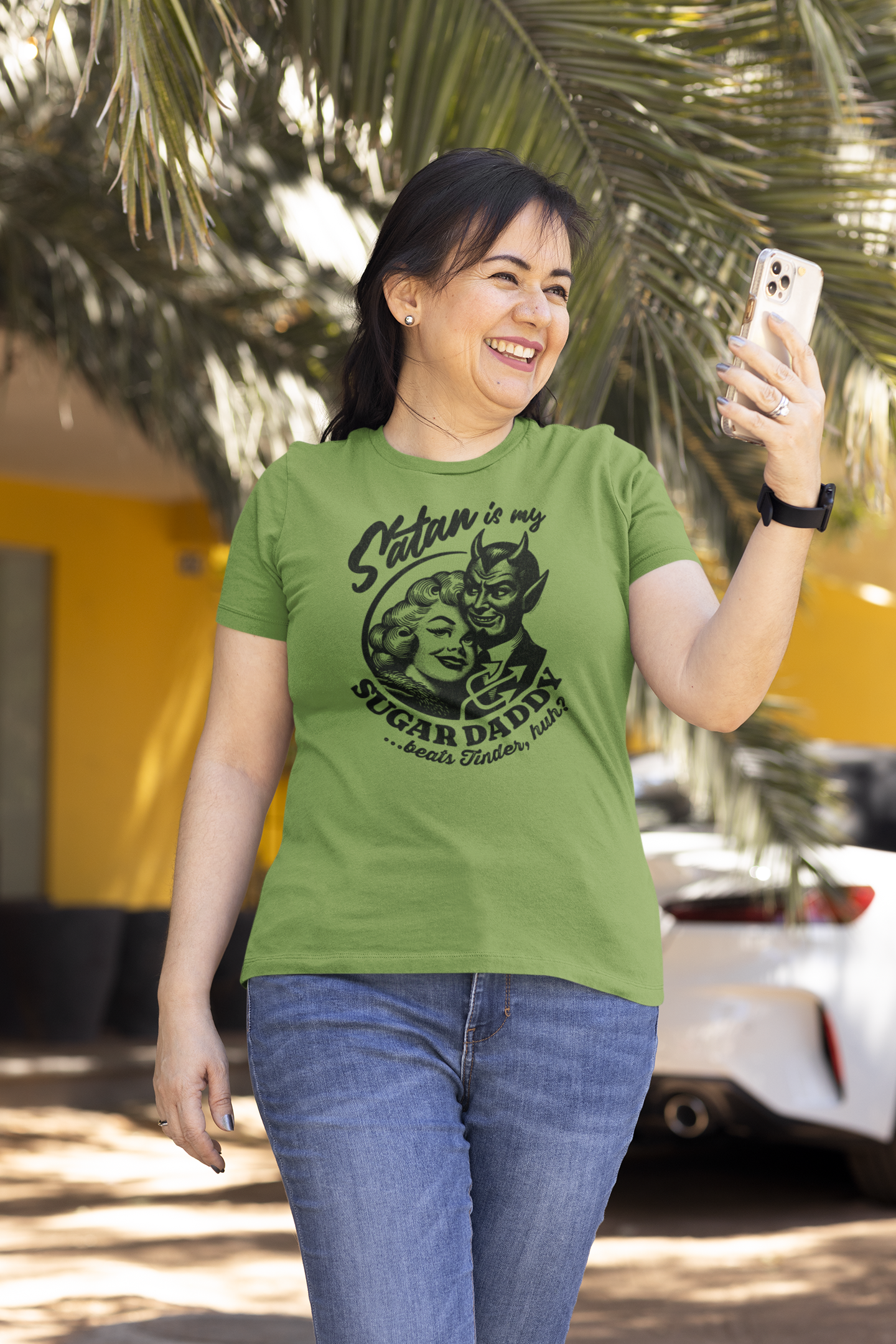 Smiling woman wearing a Leaf Green 'Satan is My Sugar Daddy' t-shirt, checking her phone while standing outside.