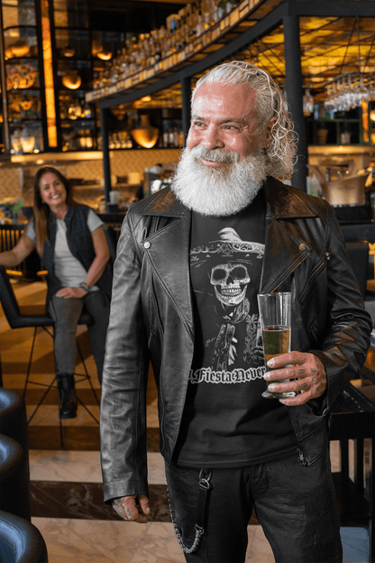 A smiling older man with a white beard wearing a black t-shirt featuring a 'La Fiesta Never Dies' Dia de los Muertos skeleton design, standing in a bar with a drink in hand.