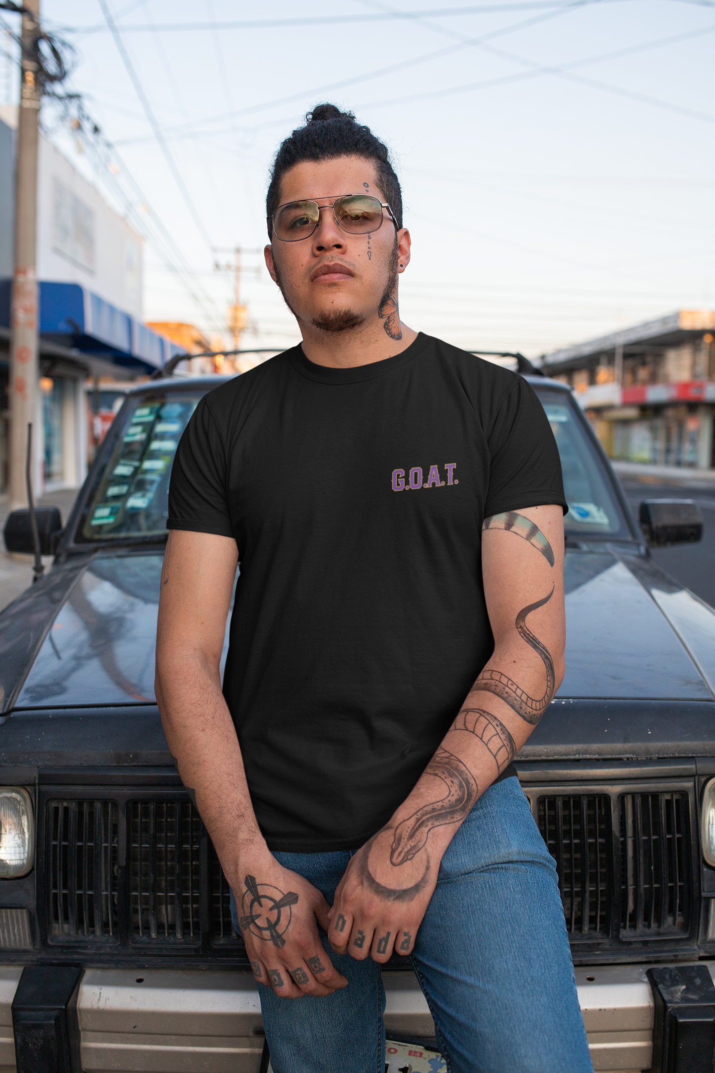 Man with tattoos posing on the street, wearing the G.O.A.T. T-Shirt in black, featuring the bold 'G.O.A.T.' text on the chest.