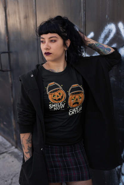 A goth woman with tattoos wearing a black t-shirt featuring the 'Smile Now, Cry Later' jack-o'-lantern design, leaning against a wall with a thoughtful expression. The retro Halloween design stands out on the t-shirt.