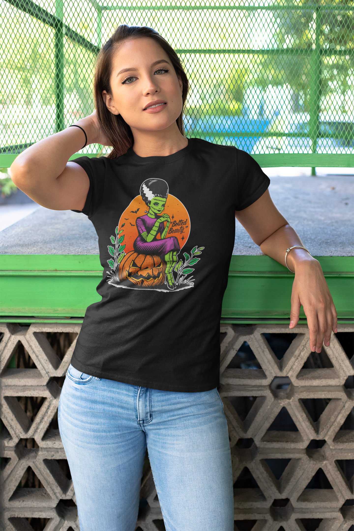 Young woman wearing a black Halloween t-shirt with a 'Bolted Beauty' design, featuring a green bride sitting on a jack-o'-lantern. She's standing by a green fence, casually posing with her hair."