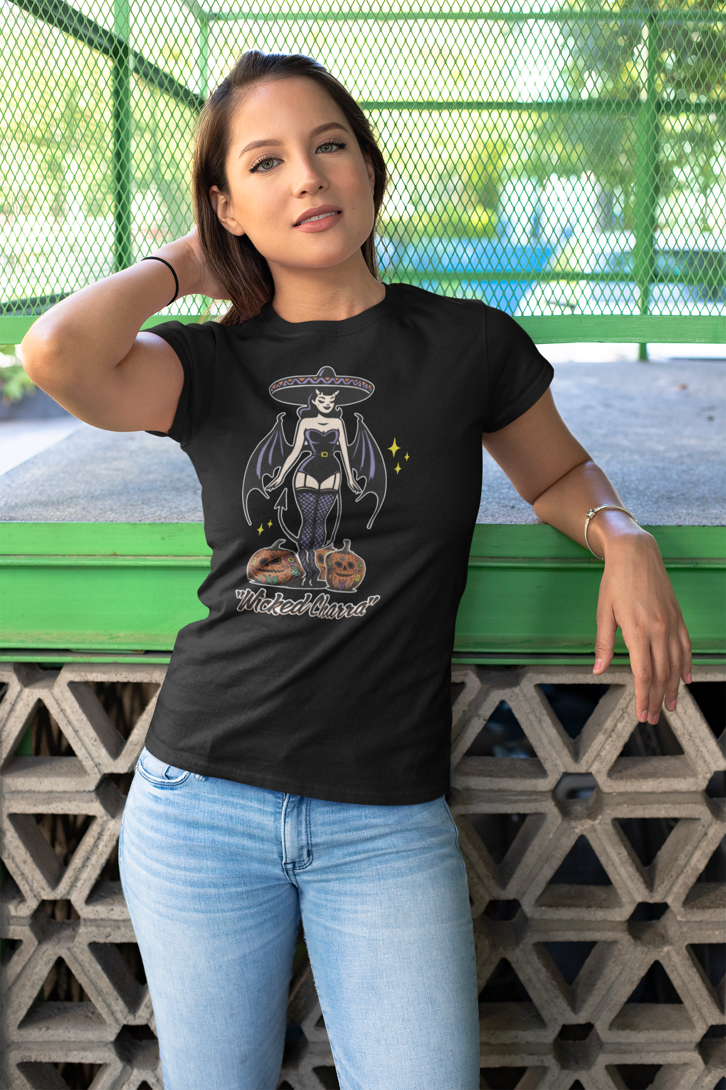 Young woman standing outdoors wearing a 'Wicked Charra' black t-shirt with a vintage-inspired charra design, featuring devil wings, a sombrero, and two jack-o-lanterns.