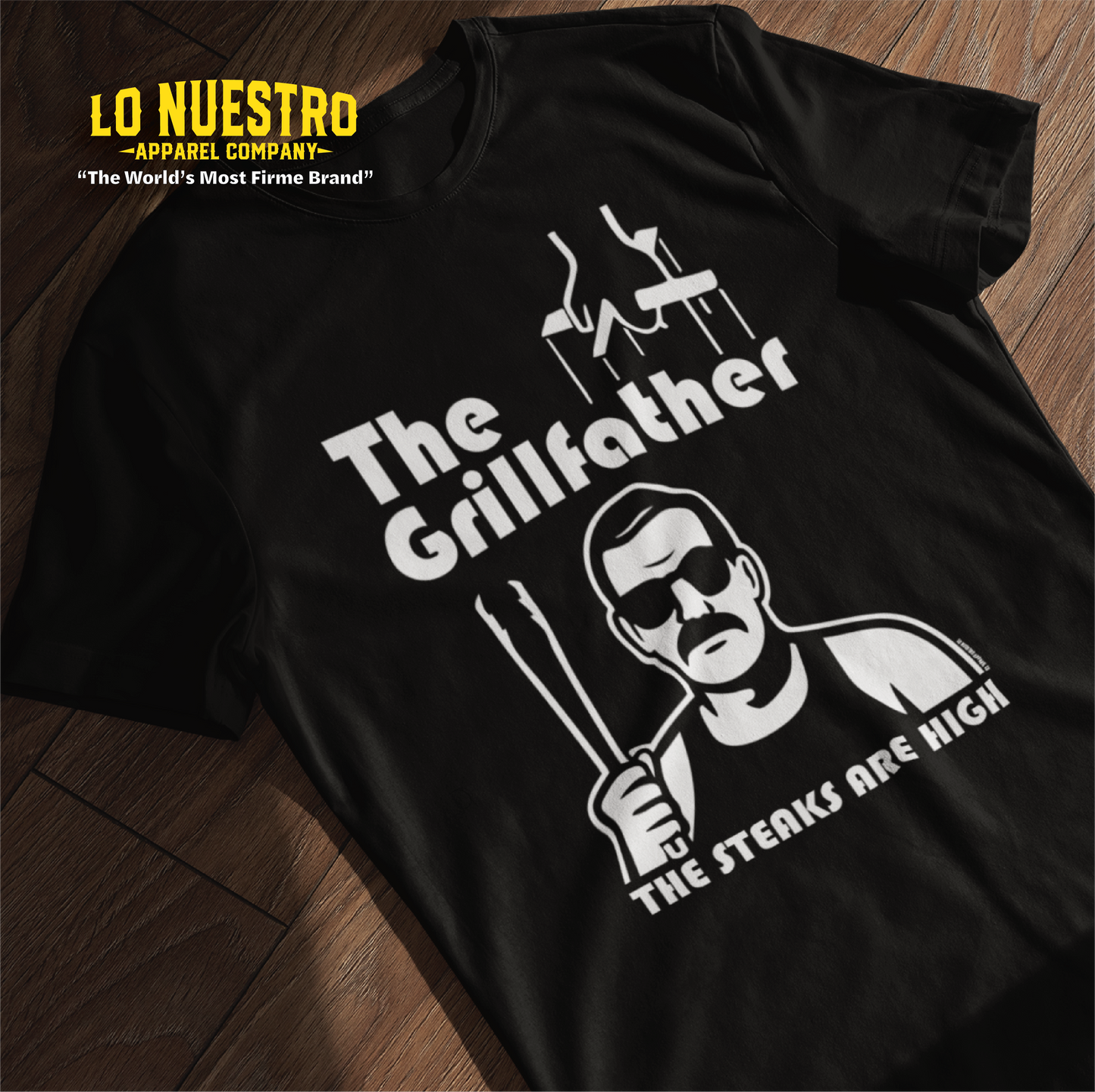 Black t-shirt laid flat featuring The Grillfather design with puppet strings, a cholo-style character holding tongs, and the phrase The Steaks Are High.