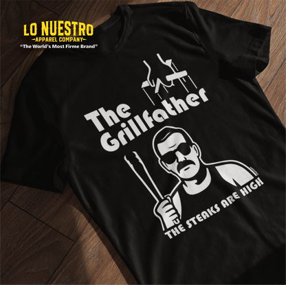 Black t-shirt laid flat featuring The Grillfather design with puppet strings, a cholo-style character holding tongs, and the phrase The Steaks Are High.