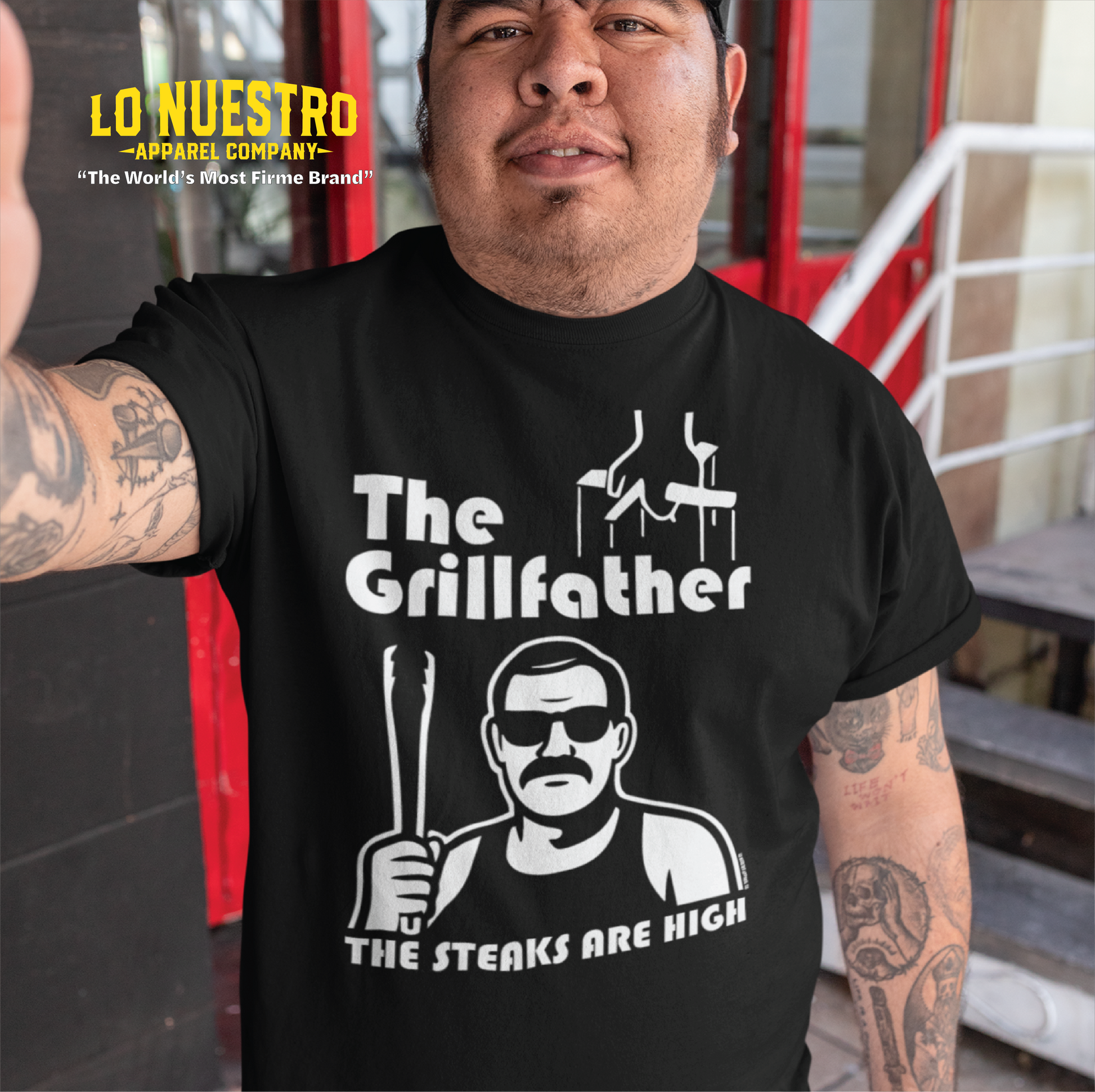 Man wearing a black t-shirt with The Grillfather design, showing puppet strings, a cholo-style grill master holding tongs, and the phrase The Steaks Are High.