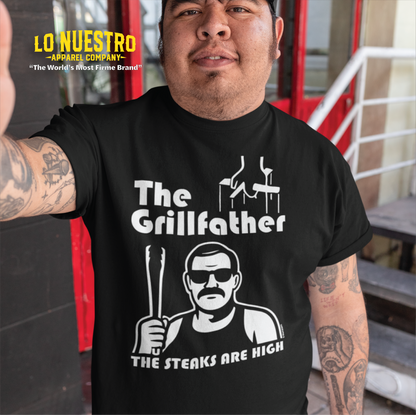Man wearing a black t-shirt with The Grillfather design, showing puppet strings, a cholo-style grill master holding tongs, and the phrase The Steaks Are High.