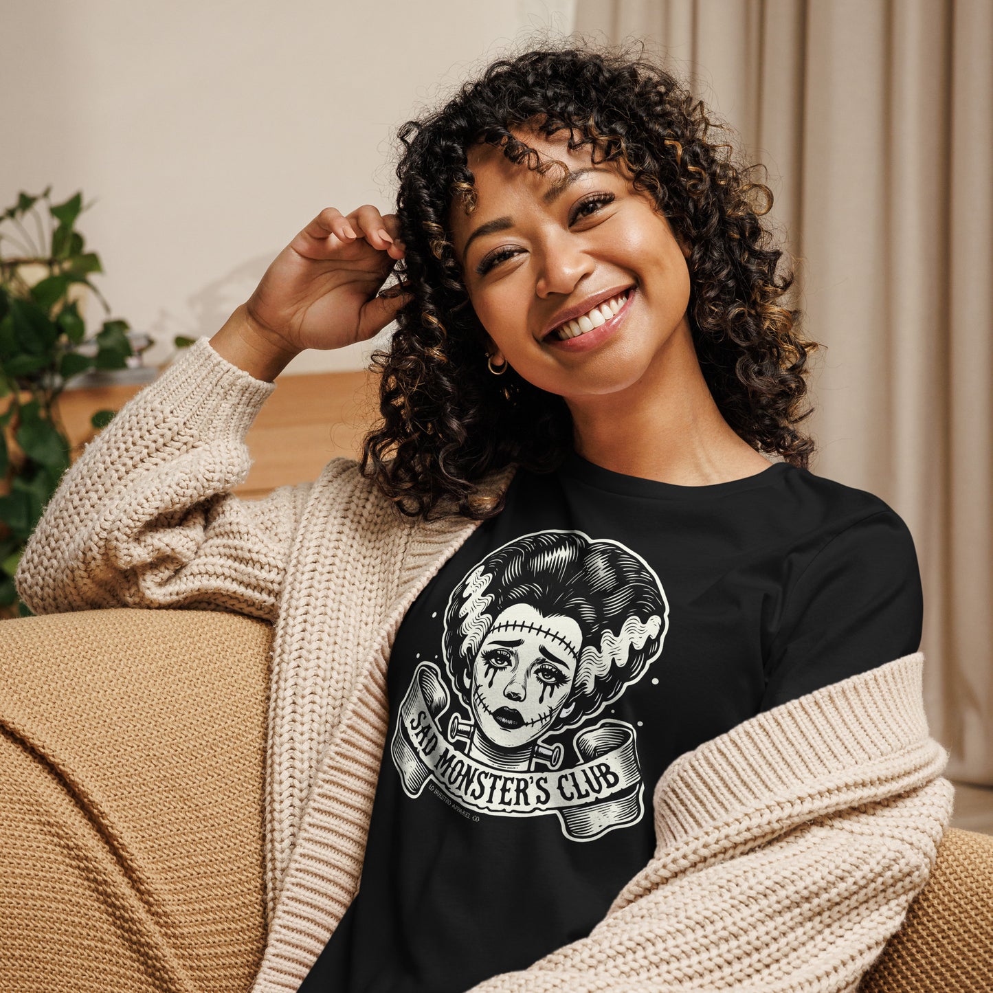 Woman smiling while wearing a black t-shirt with a retro tattoo-style Bride of Frankenstein design, featuring the 'Sad Monster's Club' banner. She is seated in a cozy living room setting with a beige cardigan over her shoulders.