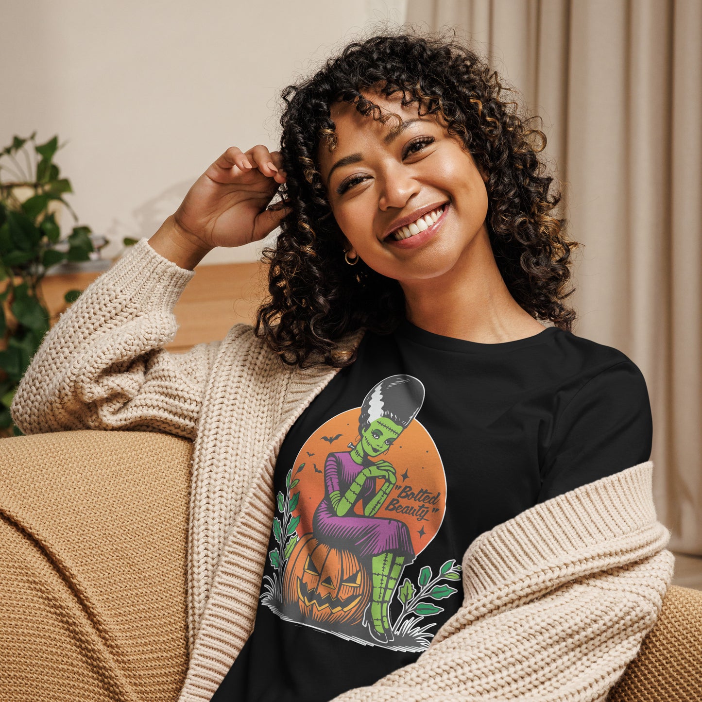 Smiling woman lounging indoors, wearing a black Halloween t-shirt with a 'Bolted Beauty' design, showing a green-skinned bride sitting on a jack-o'-lantern.